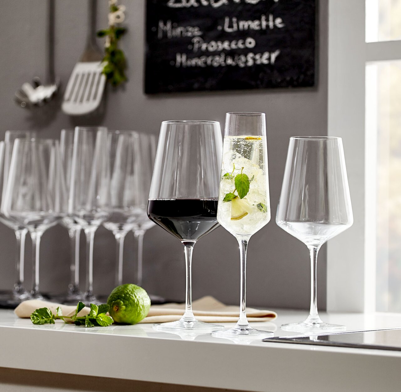 Leonardo Gläserset 18-tlg. PUCCINI Drei elegante Kelchgläser der Serie Puccini auf einer weißen Oberfläche, gefüllt mit Rotwein, einem Cocktail mit Limette und Minze, und leer, im Vordergrund mit frischen Limetten und Minzblättern, seitliche Perspektive.