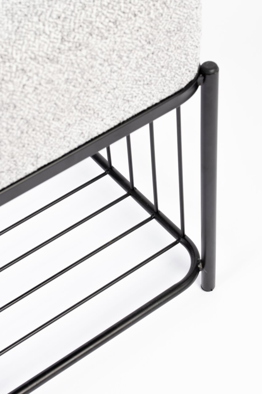 Close-up van een kruk met een zwart metalen frame en een lichtgrijze stoffen bekleding, gezien vanaf de zijkant.