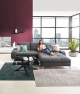 Moderner Couchtisch mit schwarzer Glasplatte und gekreuztem Metallgestell, im Vordergrund einer stilvollen Wohnzimmerumgebung mit grauem Ecksofa. Perspektive: Frontalansicht.