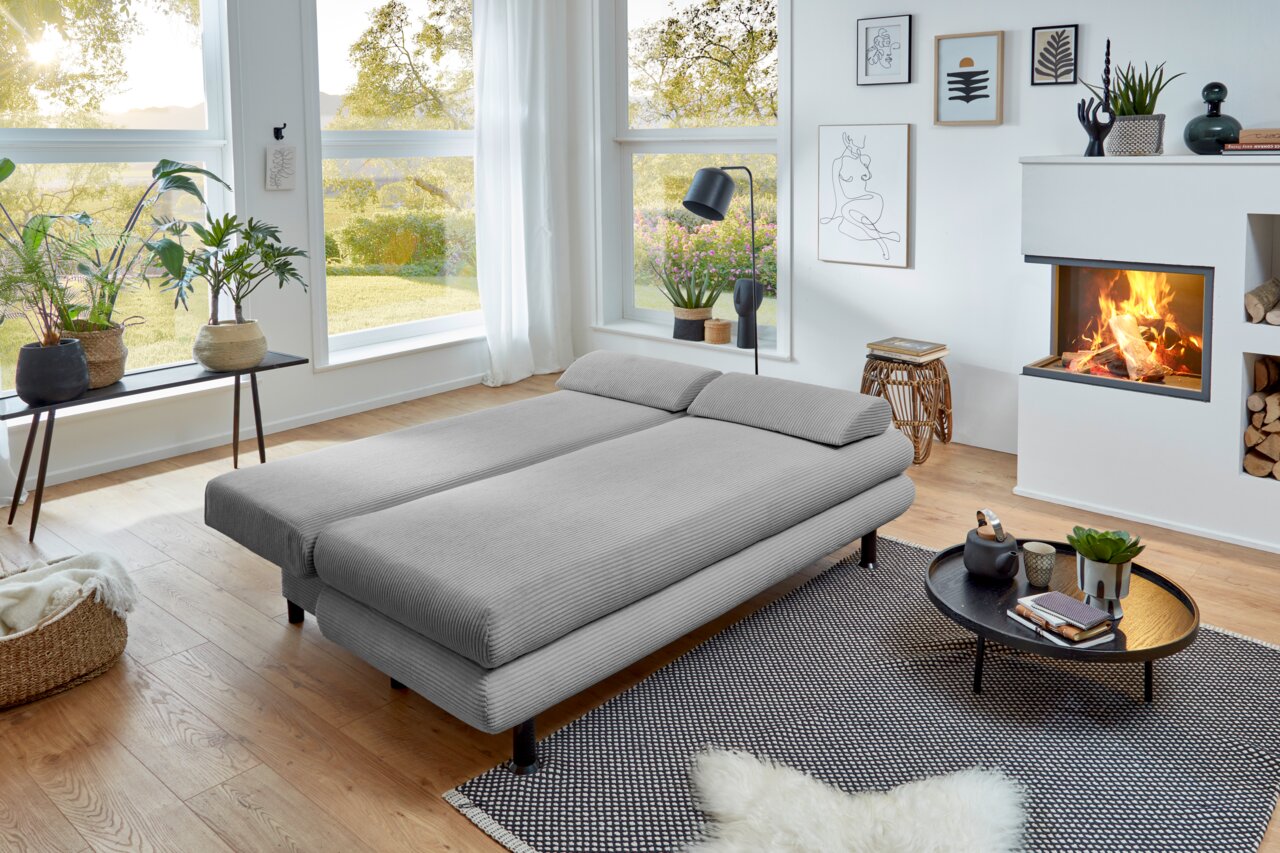 Graues Funktionssofa in einem modernen Wohnzimmer mit Blick auf den Garten, umgeben von Pflanzen und einem Kamin.