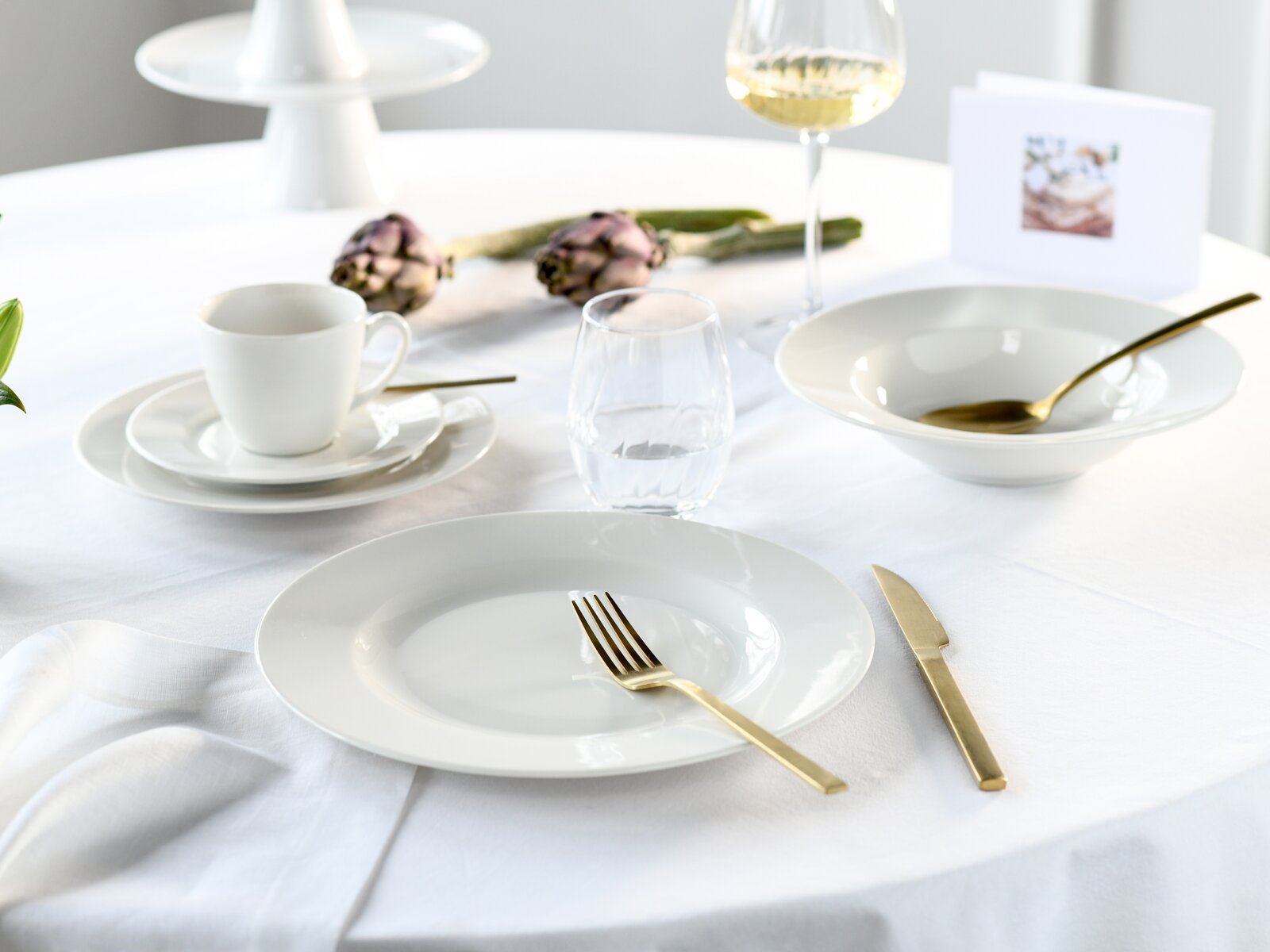 Modern gedeckter Tisch mit weißem Porzellangeschirr, goldfarbenem Besteck, Wasser- und Weinglas auf weißer Tischdecke; Perspektive: leicht seitliche Ansicht auf Tischhöhe.