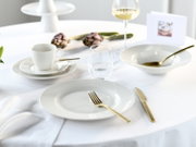 Modern gedeckter Tisch mit weißem Porzellangeschirr, goldfarbenem Besteck, Wasser- und Weinglas auf weißer Tischdecke; Perspektive: leicht seitliche Ansicht auf Tischhöhe.