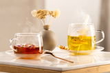Zwei Teetassen aus Glas auf einem Tisch, gefüllt mit dampfendem Tee, eine mit roten Blüten und die andere mit gelben Blüten, im Hintergrund eine Vase mit Trockenblumen, seitliche Perspektive.