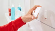 Een hand houdt een witte tesa Powerstrips-haak S recht voor een muur, zijaanzicht