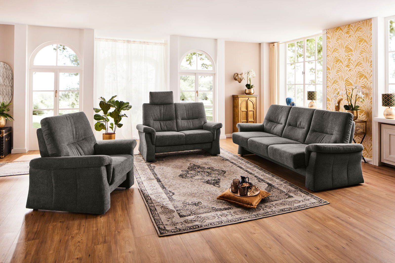 Wohnzimmeransicht mit grauem Polstermöbel-Set bestehend aus einem 3,5-Sitzer Sofa, einem 2-Sitzer Sofa und einem Sessel, auf einem Teppichboden mit Holzboden, umgeben von großen Fenstern und Pflanzen.