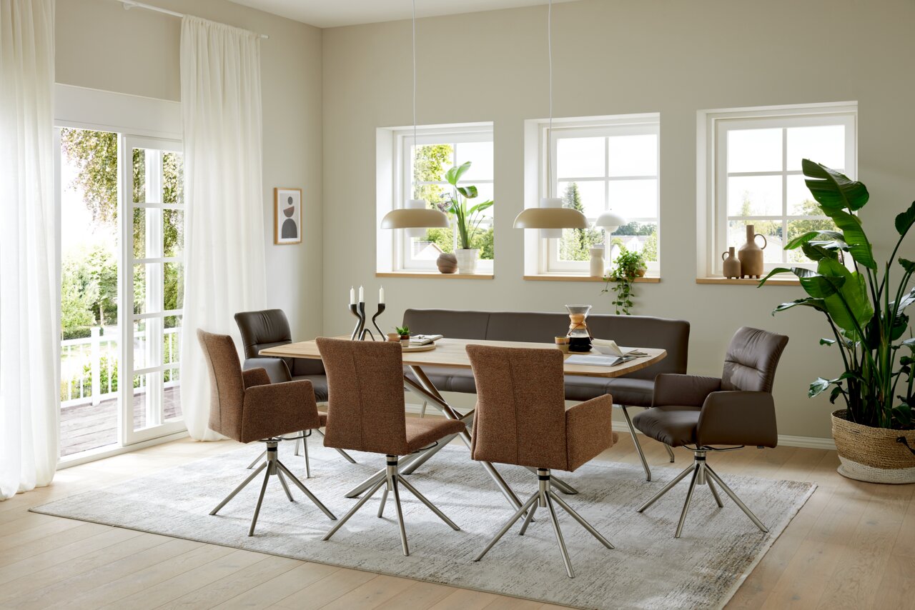 Musterring Esstisch MR HELMOND Moderner Essbereich mit einem großen Esstisch aus Holz, umgeben von stilvollen Sesseln und Stühlen in einem hellen, luftigen Raum mit großen Fenstern und Pflanzen. Perspektive von der Seite.