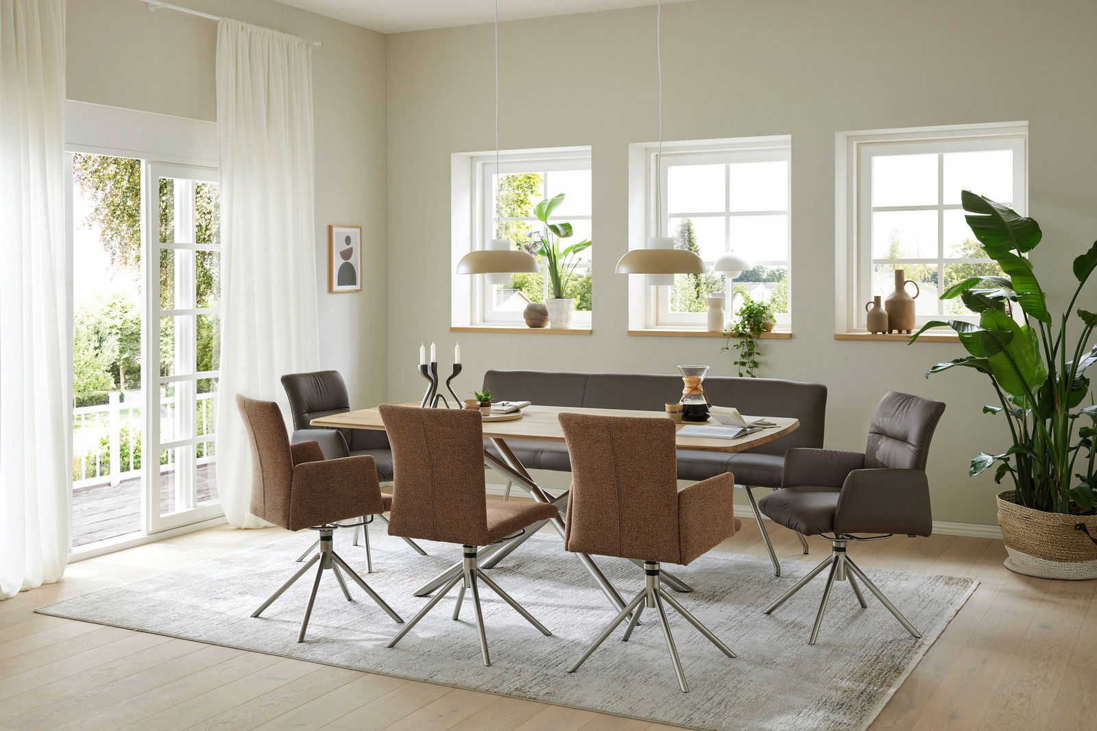 Moderner Essbereich mit einem großen Esstisch aus Holz, umgeben von stilvollen Sesseln und Stühlen in einem hellen, luftigen Raum mit großen Fenstern und Pflanzen. Perspektive von der Seite.