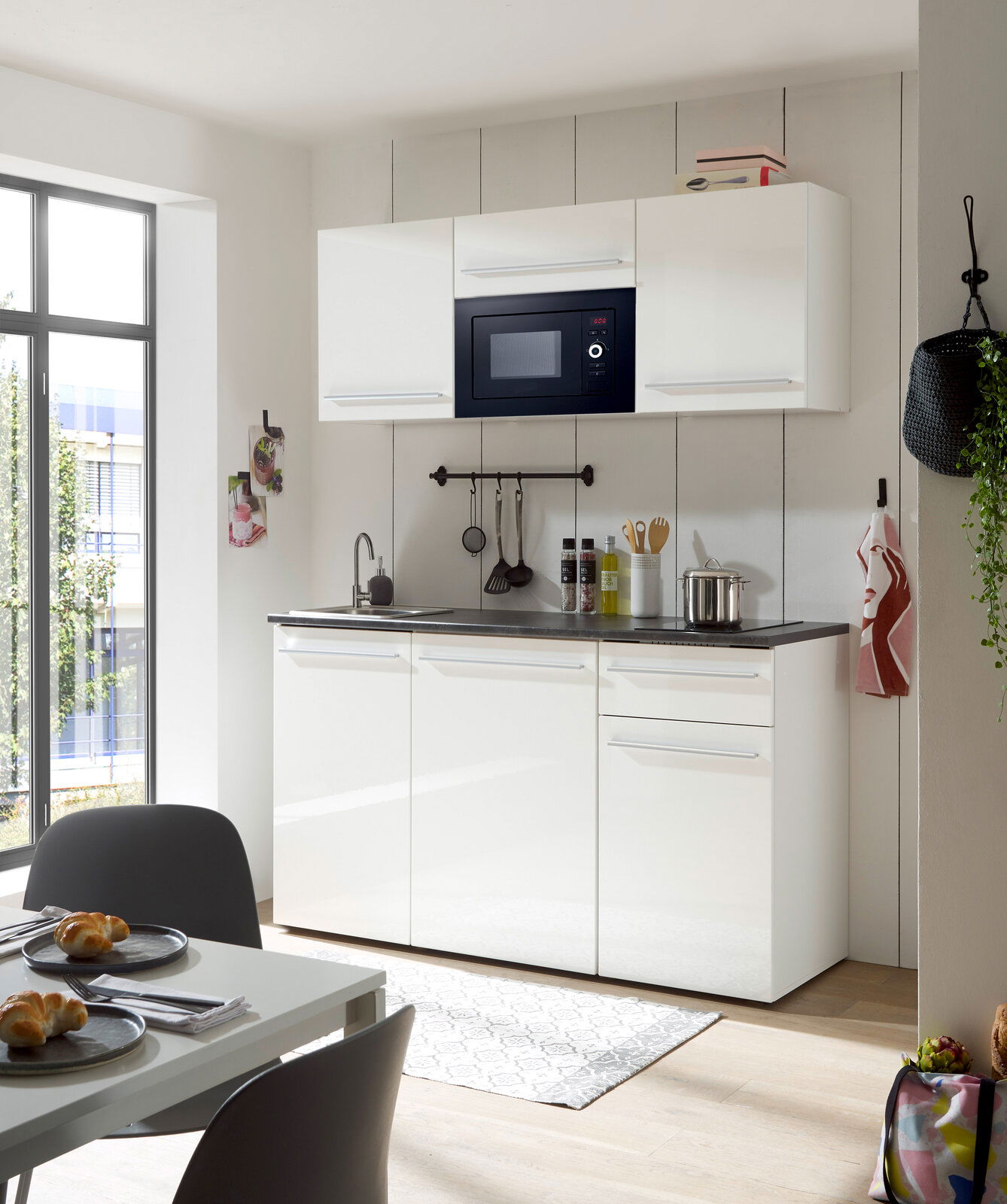 Moderne Single-Küche in Weiß mit Hängeschränken, eingebauter Mikrowelle und Spüle, aus der Perspektive eines Essbereichs mit Tisch und Stühlen.