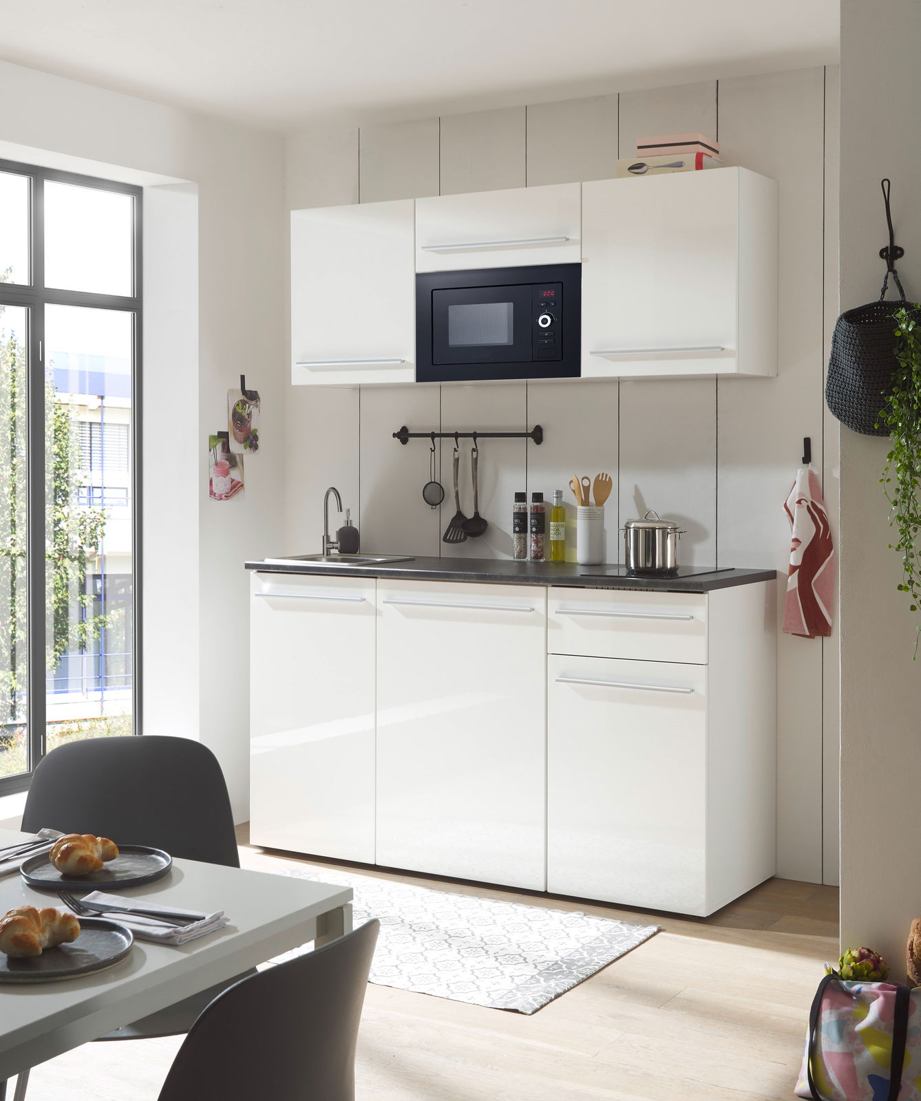 Moderne Single-Küche in Weiß mit Hängeschränken, eingebauter Mikrowelle und Spüle, aus der Perspektive eines Essbereichs mit Tisch und Stühlen.