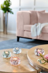Vier Teelichthalter aus Glas in verschiedenen Pastellfarben auf einem Holztisch, mit einem unscharfen Sofa im Hintergrund. Perspektive von oben.
