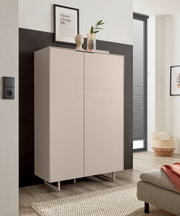 Beiger, moderner Schrank mit zwei glatten Türen und schlanken Metallfüßen in einem hellen Wohnraum, frontal aus leicht schräger Perspektive fotografiert.
