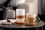 Zwei Becher mit schaumigem Kaffee auf einem Tisch, einer hoch und einer niedrig, mit einem eleganten, wellenförmigen Design, auf Korkuntersetzern, seitliche Perspektive.