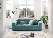 Zweiersofa in einem modernen Wohnzimmer mit großen Fenstern, grünen Pflanzen und dekorativen Kissen, Frontalansicht.