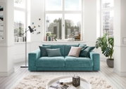 Zweiersofa in einem modernen Wohnzimmer mit großen Fenstern, grünen Pflanzen und dekorativen Kissen, Frontalansicht.