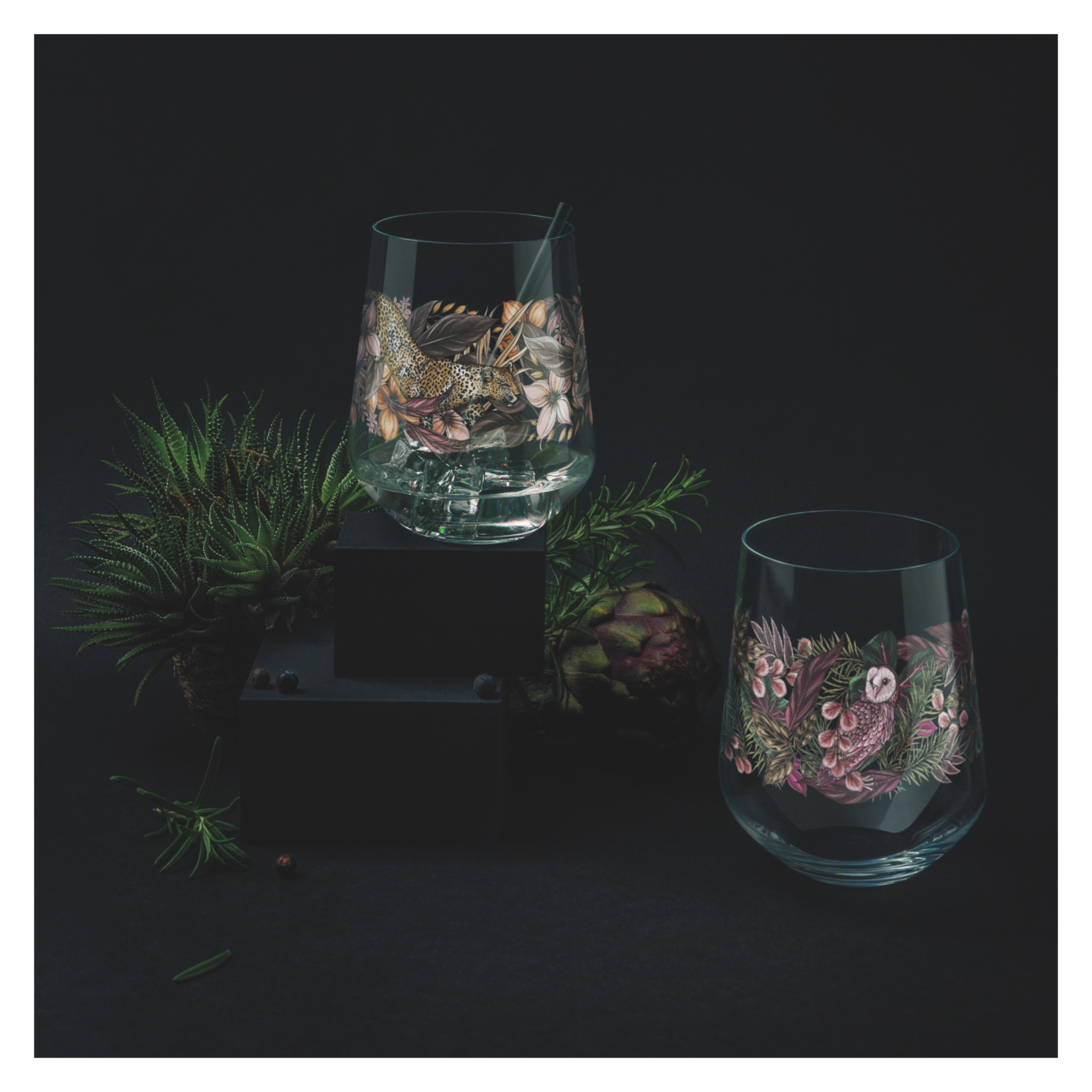 Ritzenhoff Gin Tumbler 2er-Set SCHATTENFAUNA Zwei kunstvoll verzierte Gin-Gläser in einem dunklen, eleganten Setting mit Pflanzen im Hintergrund, aus leicht erhöhter Perspektive.