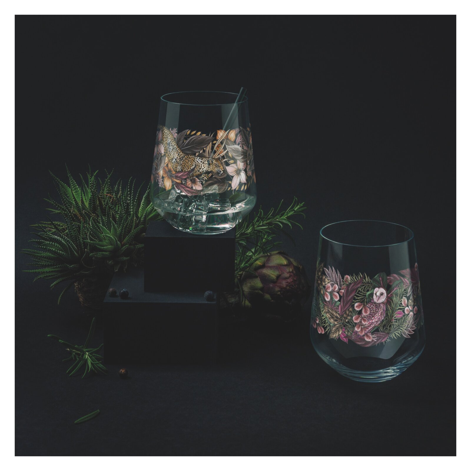 Zwei kunstvoll verzierte Gin-Gläser in einem dunklen, eleganten Setting mit Pflanzen im Hintergrund, aus leicht erhöhter Perspektive.