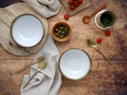 Draufsicht auf zwei weiße Keramikschalen mit rustikalem braunem Rand auf einem Holztisch, umgeben von Leinentuch, goldfarbenem Besteck, Oliven, Kirschtomaten und einem grünen Glas.