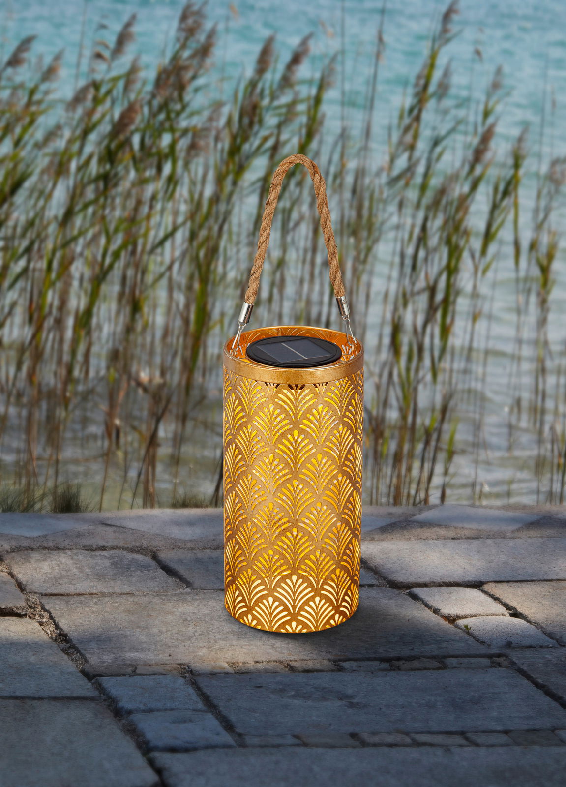 Solarbetriebene Laterne mit dekorativem Muster und Tragegriff, auf einer Terrasse mit Blick auf Schilf und Wasser.