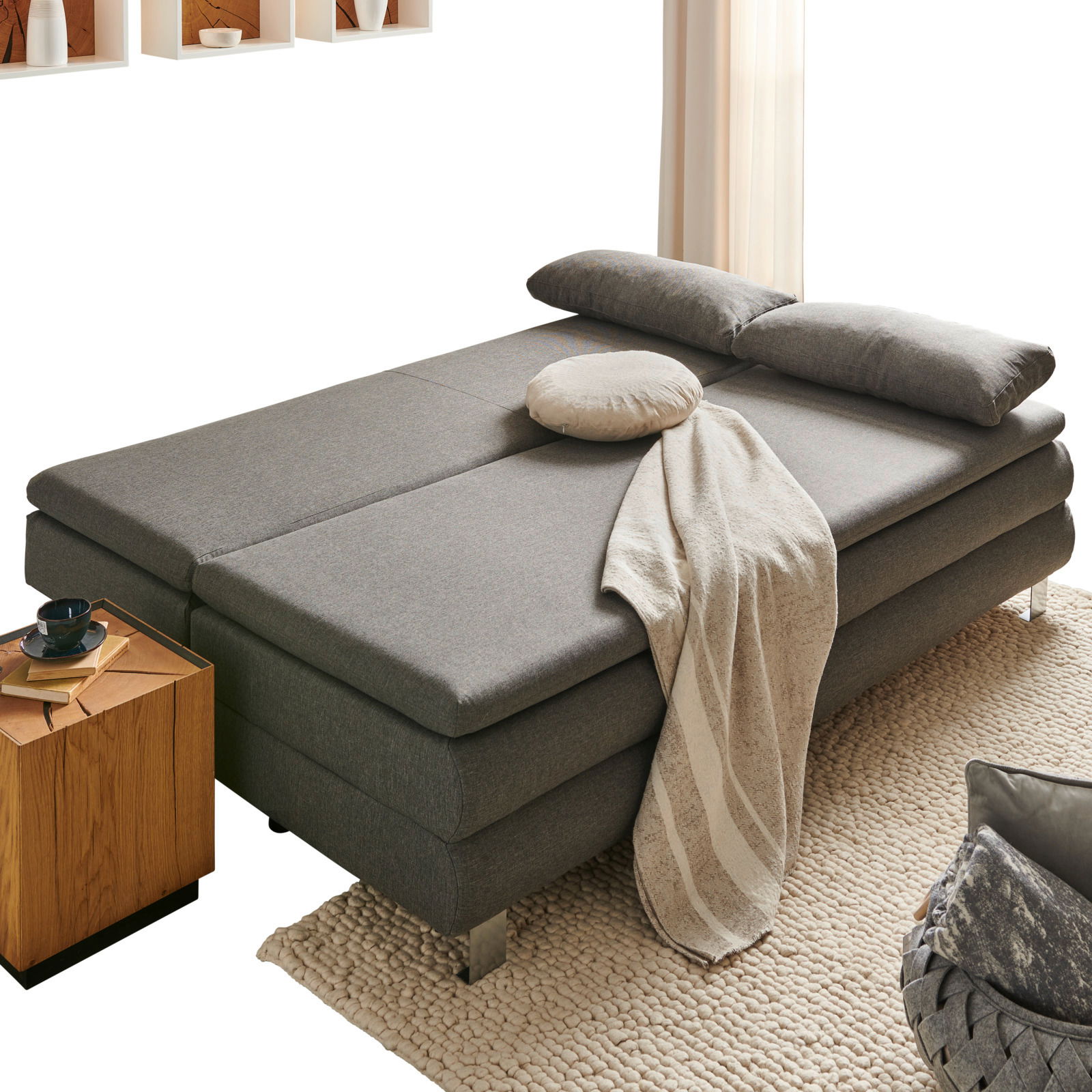 Z2 Schlafsofa GENF Graues Funktionssofa in Bettposition, seitliche Perspektive, mit Kissen und Decke dekoriert, neben einem Holztisch auf einem Teppich.