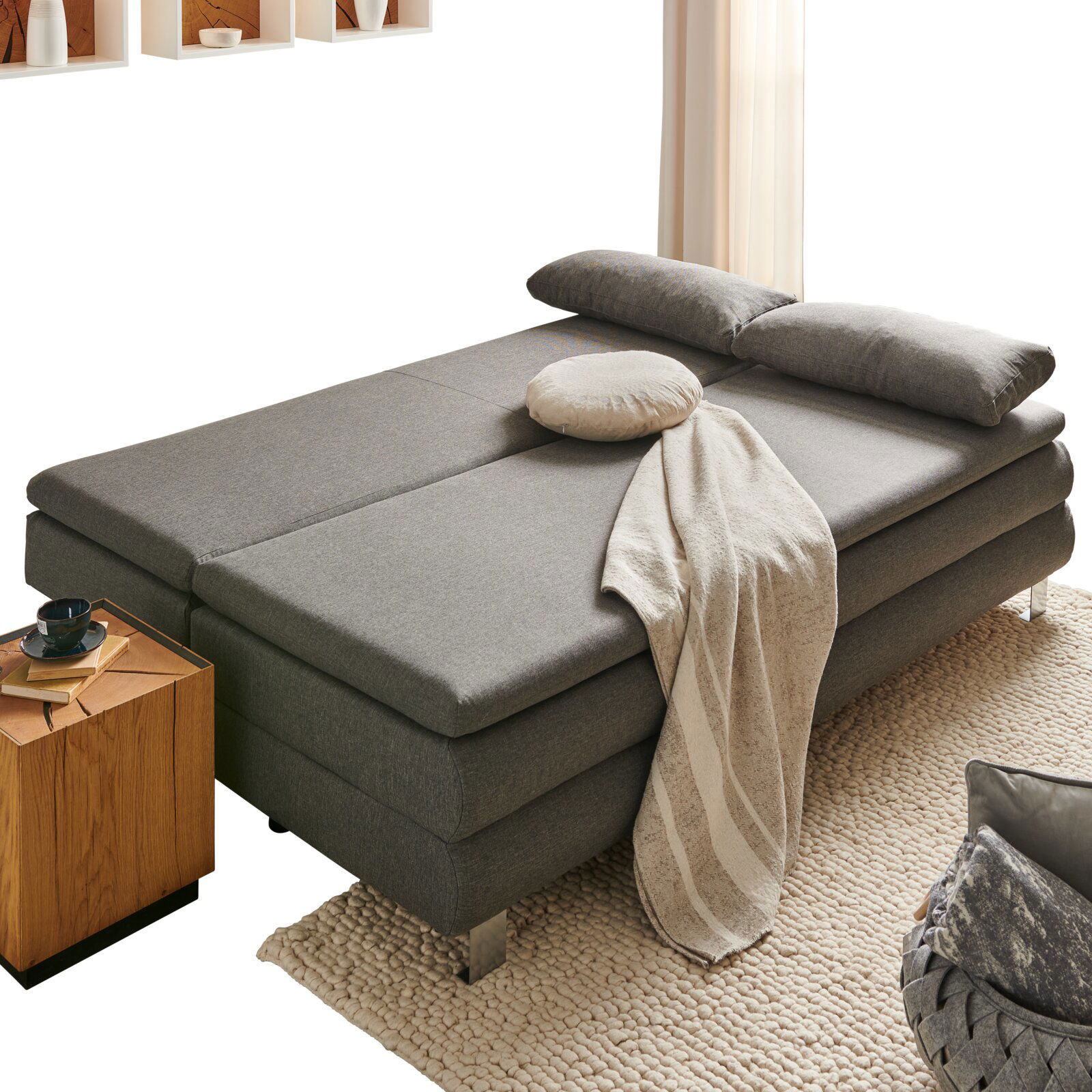 Z2 Schlafsofa GENF Graues Funktionssofa in Bettposition, seitliche Perspektive, mit Kissen und Decke dekoriert, neben einem Holztisch auf einem Teppich.