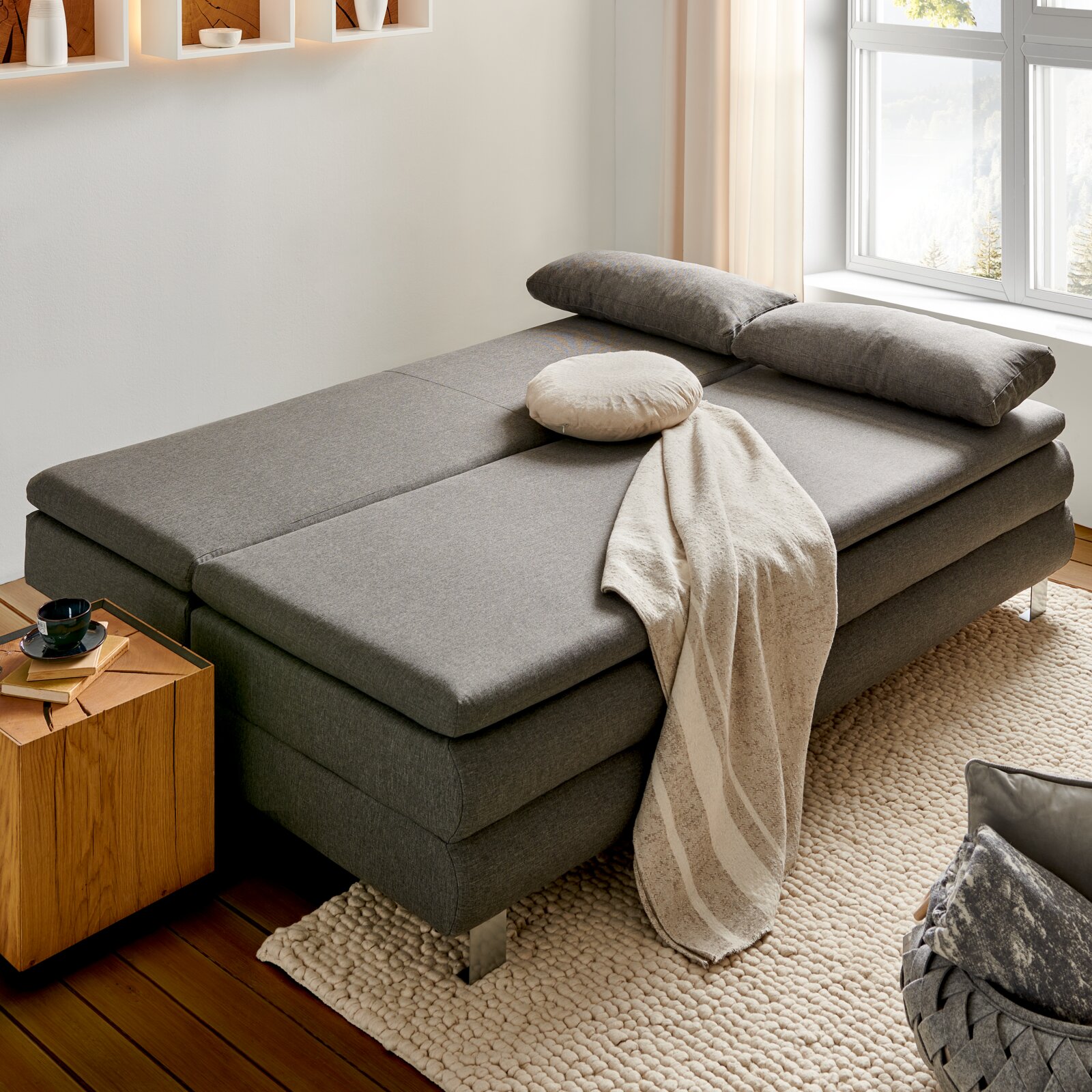 Z2 Schlafsofa GENF Graues Funktionssofa in Bettposition, seitliche Perspektive, mit Kissen und Decke dekoriert, neben einem Holztisch auf einem Teppich.
