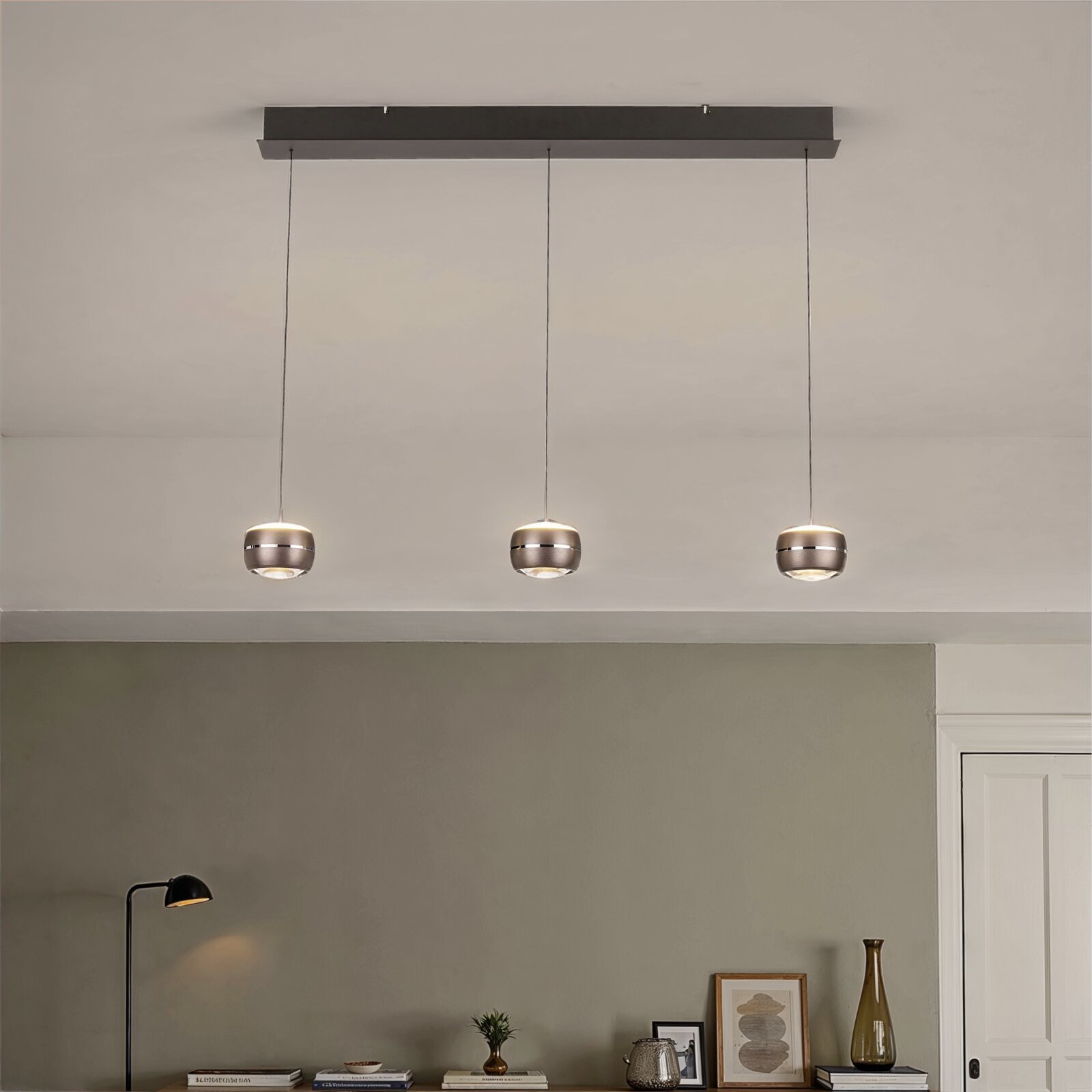 Moderne hanglamp met drie lampen, voorzien van drie ronde metalen kappen aan lange kabels en een rechthoekige plafondbeugel, gezien vanaf de voorkant vanuit een licht naar beneden gericht perspectief in de woonkamer