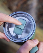 Close-up van bovenaf van een transparant drinkbekerdeksel met blauwgroene schuifsluiting die met een vinger wordt bediend