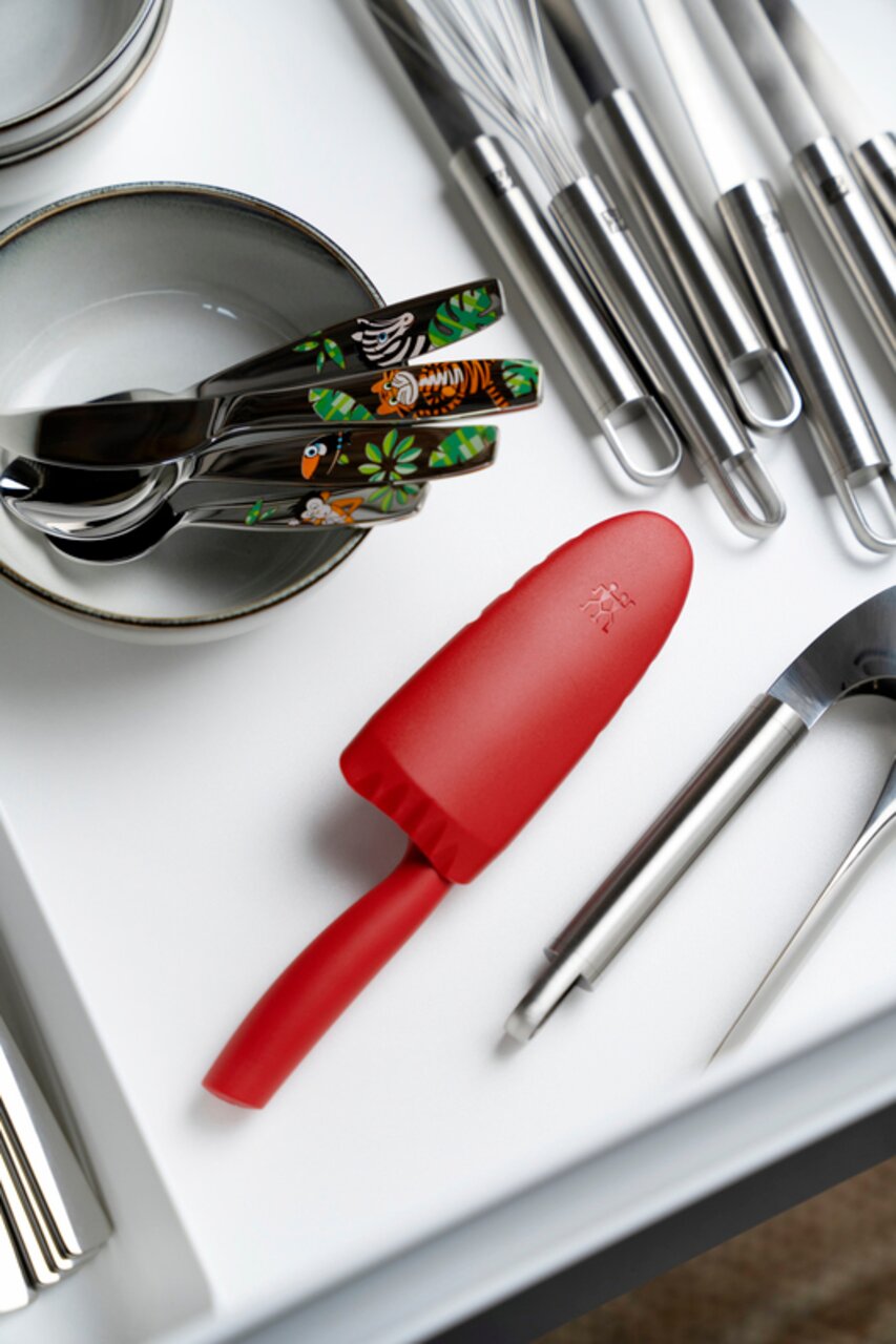 Rotes Kinderkochmesser mit 10 cm Klinge, liegend in einer Schublade neben Besteck und Küchenutensilien, aus der Vogelperspektive.