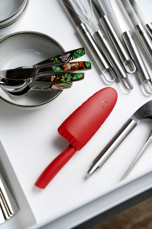 Rotes Kinderkochmesser mit 10 cm Klinge, liegend in einer Schublade neben Besteck und Küchenutensilien, aus der Vogelperspektive.