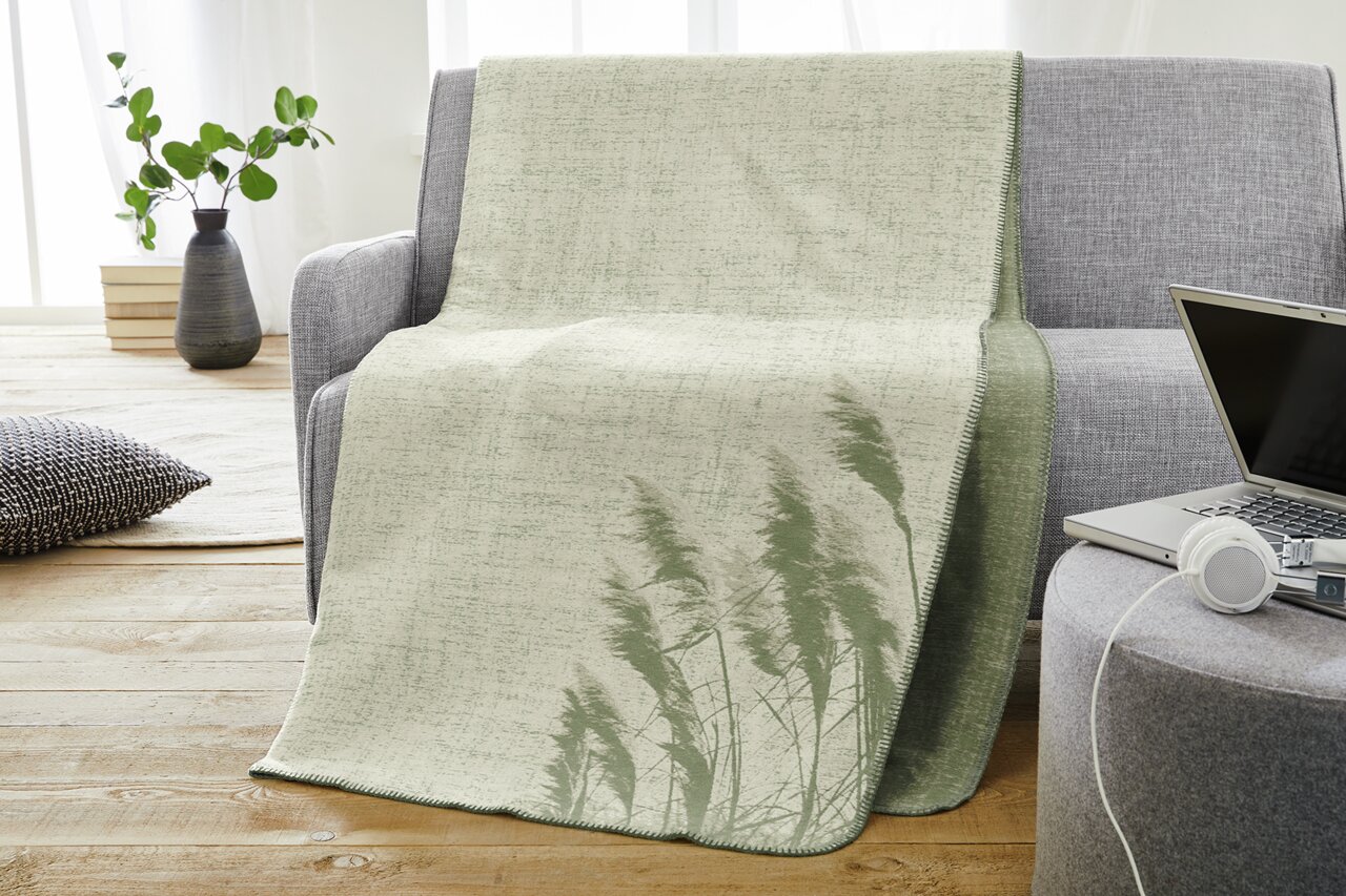 Jacquard Decke in Grün- und Beigetönen mit Pflanzenmuster, drapiert über einem grauen Sofa in einem modernen Wohnzimmer.