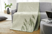 Jacquard Decke in Grün- und Beigetönen mit Pflanzenmuster, drapiert über einem grauen Sofa in einem modernen Wohnzimmer.