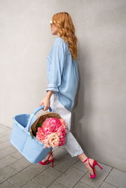 Frau in blauer Jeansbluse und weißen Hosen trägt einen hellblauen Einkaufskorb mit Blumen, seitliche Perspektive