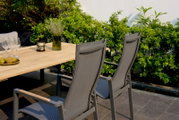 Twee grijze stapelstoelen van metaal en textiel, van achteren gefotografeerd, staan bij een houten tafel in de tuin, omringd door groene planten.
