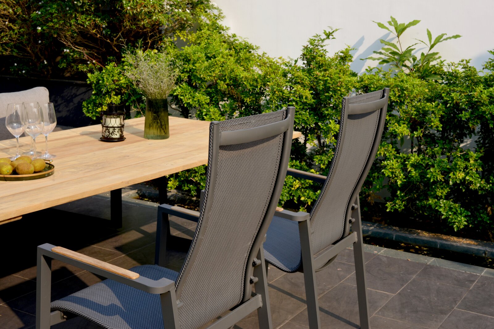 Twee grijze stapelstoelen van metaal en textiel, van achteren gefotografeerd, staan bij een houten tafel in de tuin, omringd door groene planten.