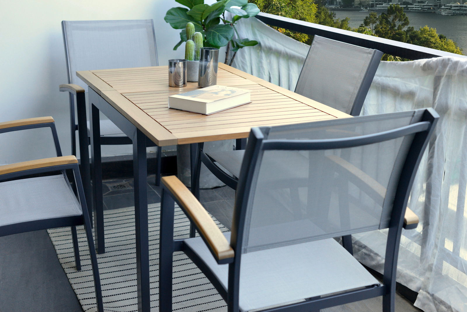 Moderner Gartentisch mit Holzplatte und Metallgestell, umgeben von Stapelsesseln auf einem Balkon mit Blick auf die Umgebung, seitliche Perspektive.