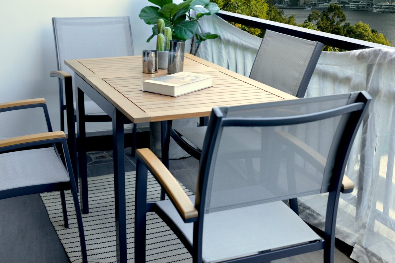 Moderner Gartentisch mit Holzplatte und Metallgestell, umgeben von Stapelsesseln auf einem Balkon mit Blick auf die Umgebung, seitliche Perspektive.