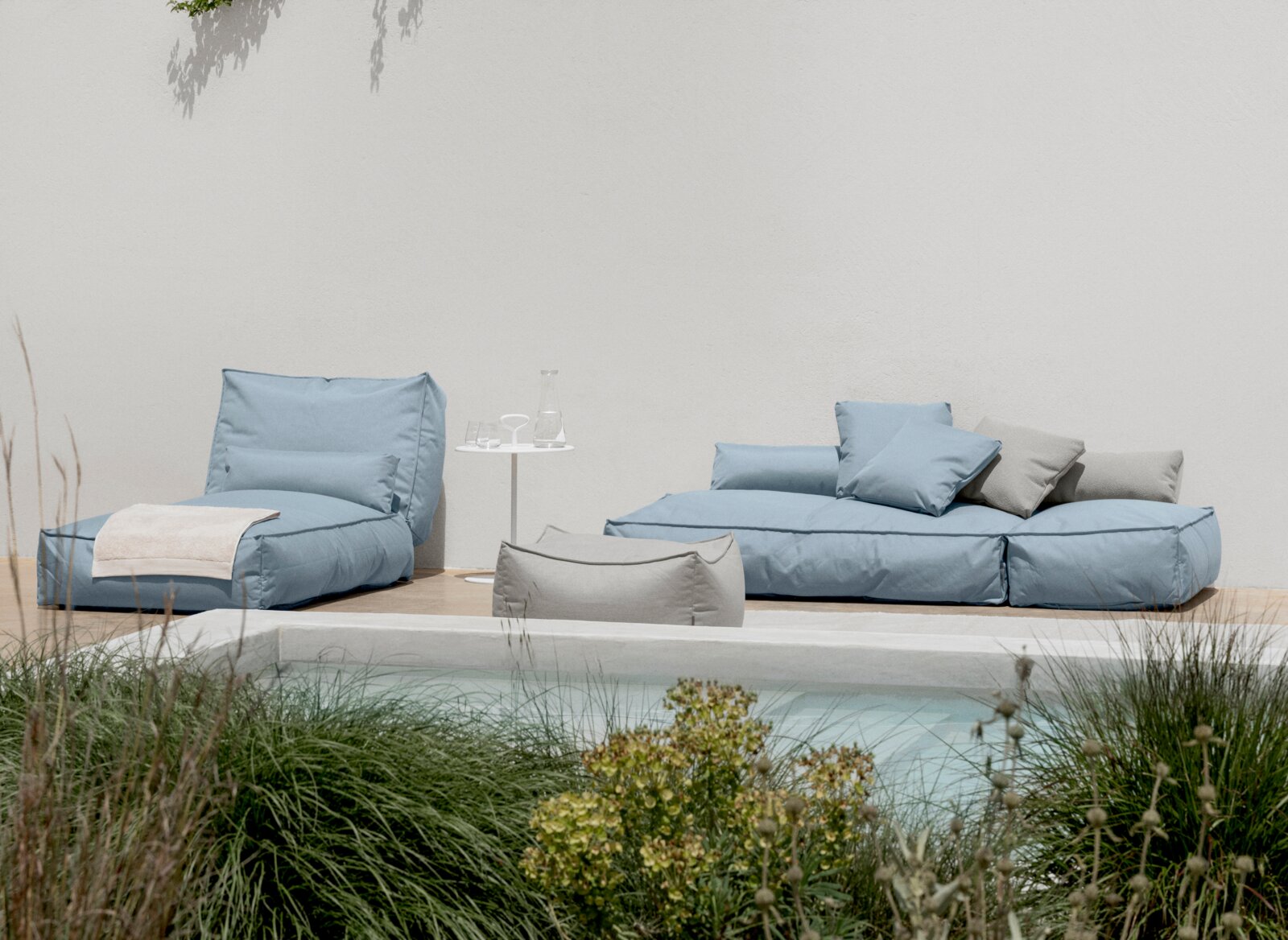 Outdoor-Lounge-Set mit blau gepolstertem Sessel und modularer Sofa-Liege neben einem Pool, mit grauen Kissen und kleinem weißem Beistelltisch; Perspektive: frontale, leicht erhöhte Ansicht.