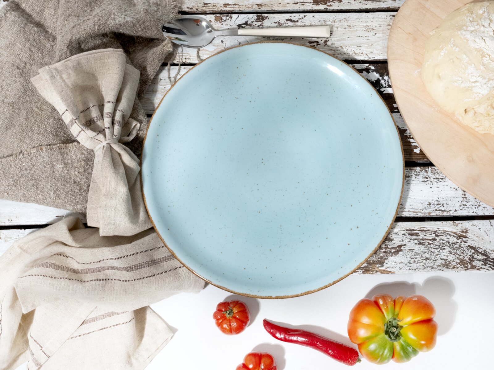 Hellblauer, gesprenkelter Keramikteller mit braunem Rand auf rustikalem Holztisch, Draufsicht; daneben Stoffserviette, Löffel, Teig auf Holzbrett sowie Tomaten und rote Chili