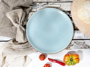 Hellblauer, gesprenkelter Keramikteller mit braunem Rand auf rustikalem Holztisch, Draufsicht; daneben Stoffserviette, Löffel, Teig auf Holzbrett sowie Tomaten und rote Chili