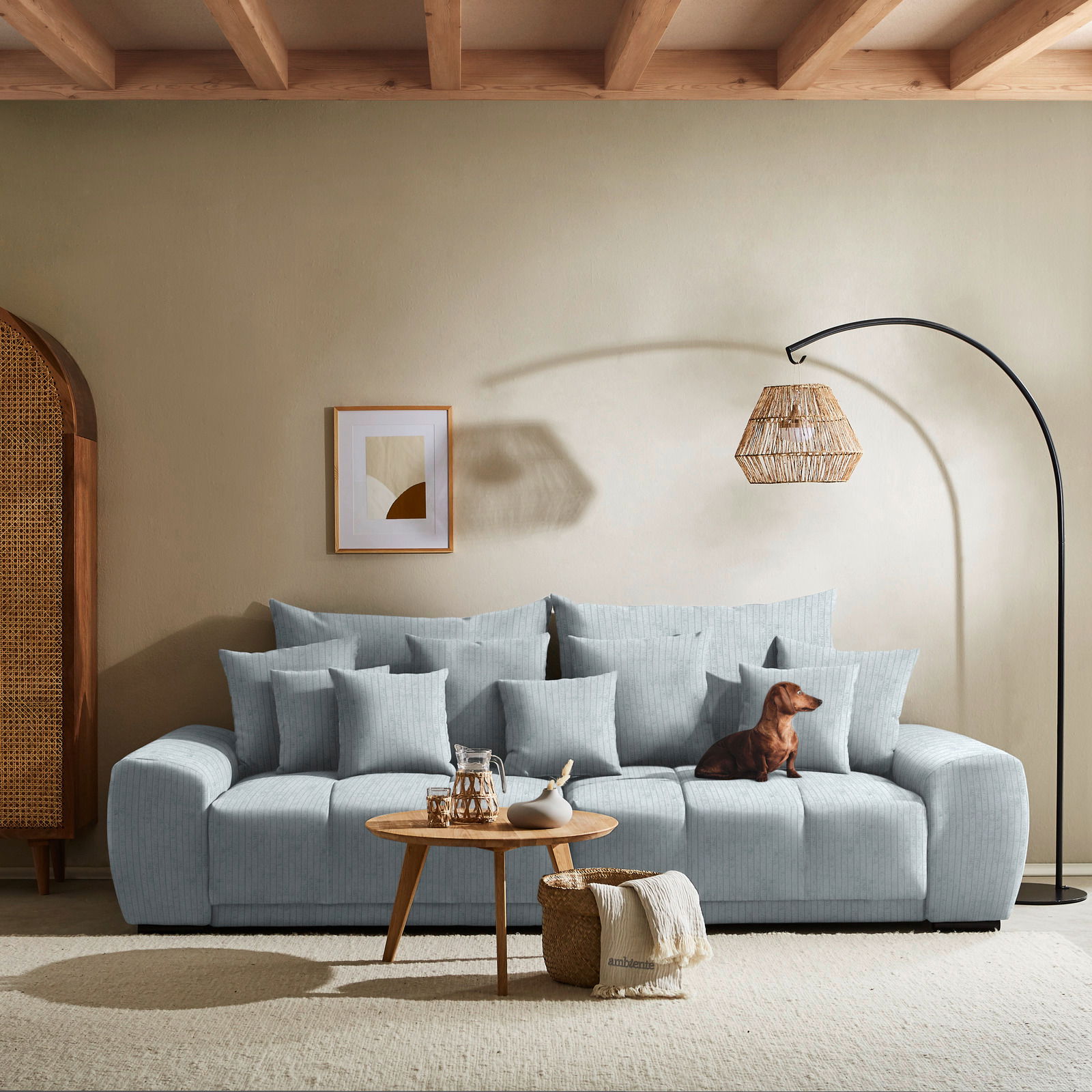 Großes, hellblaues Sofa mit vielen Kissen, ein kleiner Hund sitzt darauf. Im Vordergrund ein runder Holztisch mit Dekoration. Perspektive: Frontalansicht.