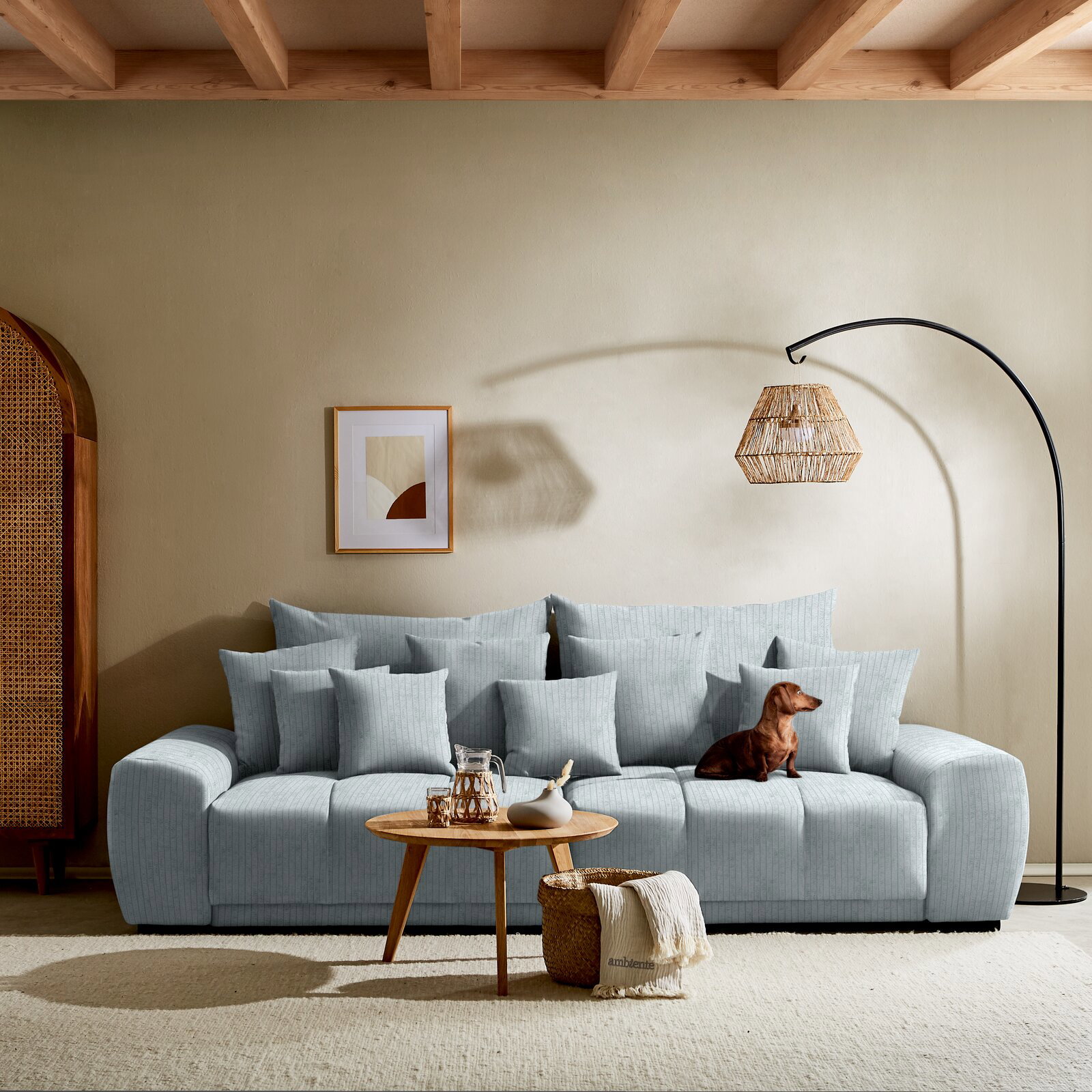 Großes, hellblaues Sofa mit vielen Kissen, ein kleiner Hund sitzt darauf. Im Vordergrund ein runder Holztisch mit Dekoration. Perspektive: Frontalansicht.