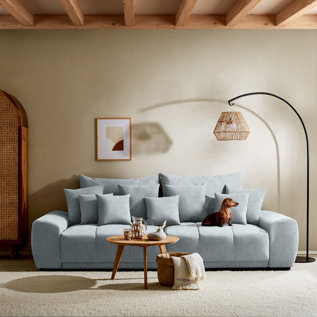 Großes, hellblaues Sofa mit vielen Kissen, ein kleiner Hund sitzt darauf. Im Vordergrund ein runder Holztisch mit Dekoration. Perspektive: Frontalansicht.