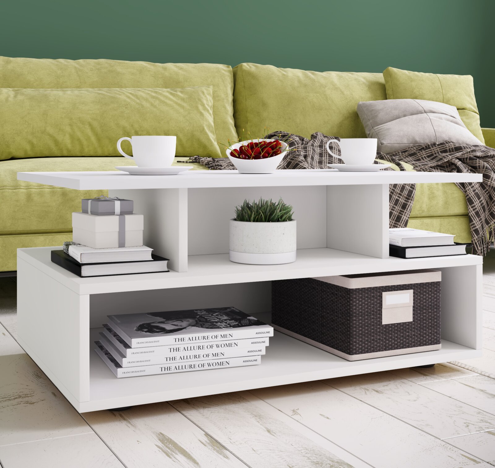Weißer, moderner Couchtisch mit Ablagefächern vor grünem Sofa, dekoriert mit zwei Tassen, Schale mit Kirschen, Büchern, Pflanze und Korb, in frontaler leicht erhöhter Perspektive.