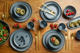 Graues Kombiservice Urban 16-teilig auf einem Holztisch, bestehend aus Tellern, Schalen und Tassen, mit goldfarbenem Besteck und frischen Zutaten wie Tomaten und Blaubeeren. Draufsicht.
