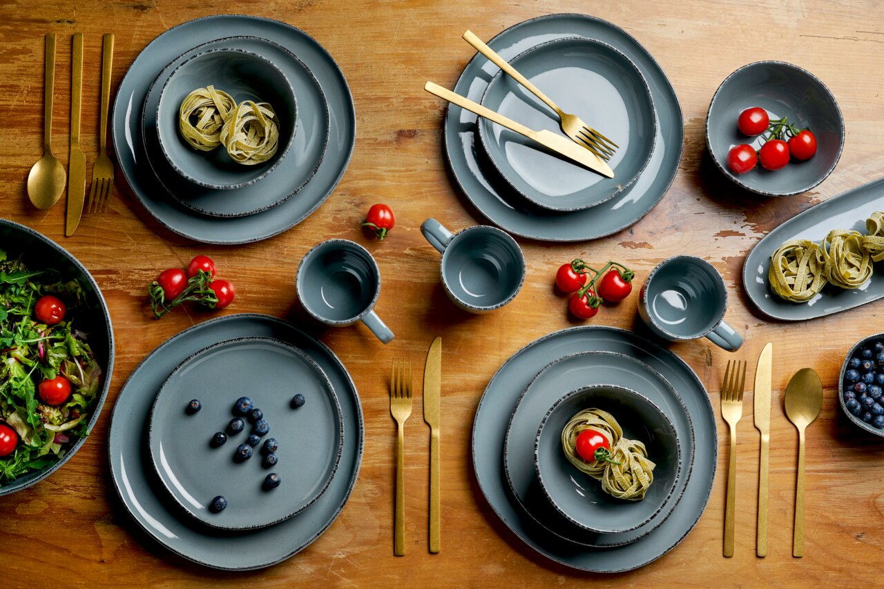 Graues Kombiservice Urban 16-teilig auf einem Holztisch, bestehend aus Tellern, Schalen und Tassen, mit goldfarbenem Besteck und frischen Zutaten wie Tomaten und Blaubeeren. Draufsicht.