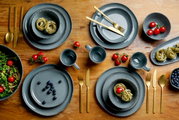 Graues Kombiservice Urban 16-teilig auf einem Holztisch, bestehend aus Tellern, Schalen und Tassen, mit goldfarbenem Besteck und frischen Zutaten wie Tomaten und Blaubeeren. Draufsicht.