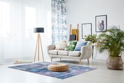 Moderner Wohnraum mit einem blauen Teppich in abstraktem Design, einem hellen Sofa mit bunten Kissen und einer Stehlampe, aus der Perspektive von vorne.
