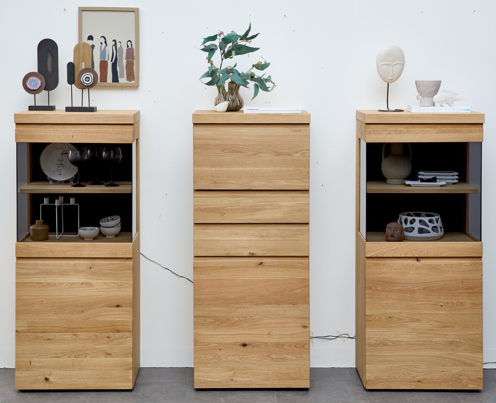 Drei moderne Hochschränke aus naturfarbener Eiche mit Schubladen und teils offenen, dunkel hinterlegten Regalfächern, dekoriert mit Vasen und Wohnaccessoires, frontal dargestellt.
