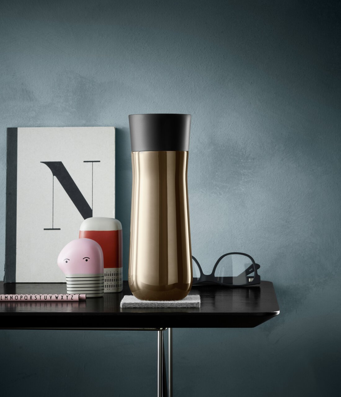 Goldener Isolierbecher mit schwarzem Deckel, frontal auf einem Tisch vor einer blauen Wand platziert.