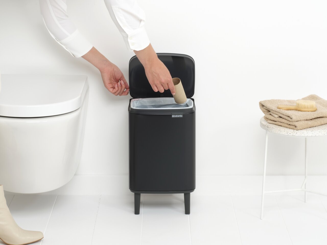 Brabantia Abfallbehälter Schwarzer Abfalleimer Bo Hi 7L mit geöffnetem Deckel, von vorne gesehen, neben einer Toilette und einem Tisch mit Handtüchern.